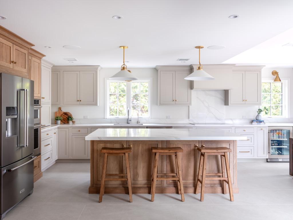 Bogert Home kitchen — greige shaker cabinetry with a white oak island, three wooden counter stools, twin white pendant lights and a marble slab behind the range