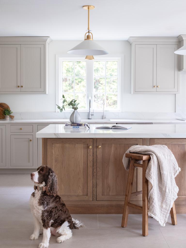 Bogert Home kitchen — marble-topped island sink under a brass and white pendant, with a linen throw over a wooden stool and a springer spaniel on the tiled floor