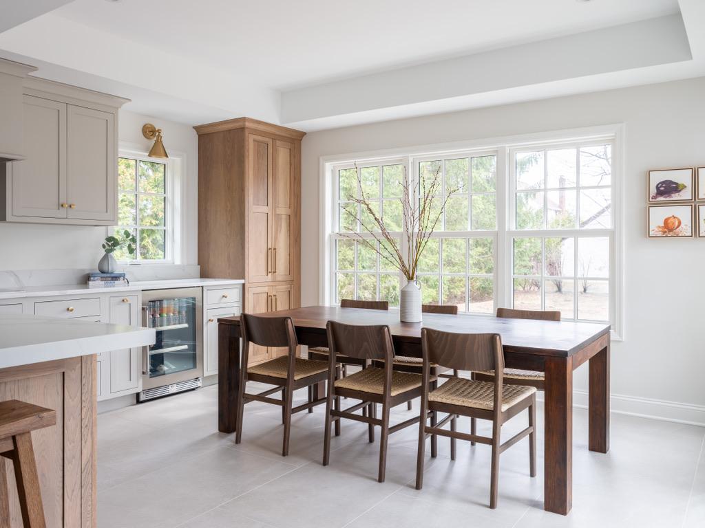 Bogert Home dining area — walnut table and woven-seat chairs beneath a bank of windows, alongside a white oak pantry cabinet and glass-fronted beverage fridge
