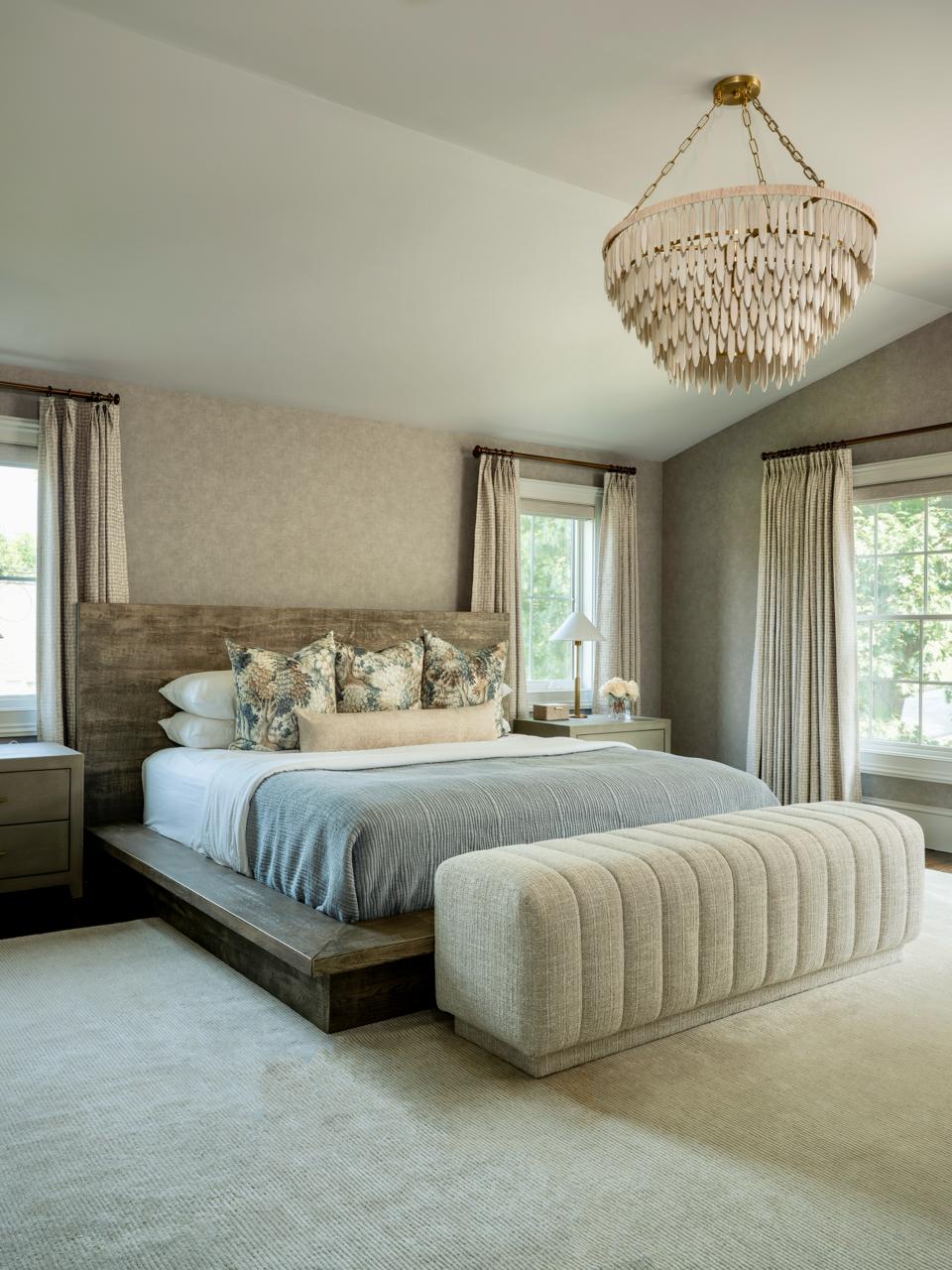 Serene primary bedroom — a rustic wood platform bed with a blue-grey coverlet and botanical pillows, a channel-tufted cream bench at the foot, and a shell chandelier above